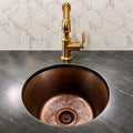 Round hammered antique copper bar sink undermounted in a black marble counter, front view with brass faucet