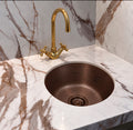 Round hammered antique copper bar sink in a marble corner bar with a brass cross handle faucet and matching strainer
