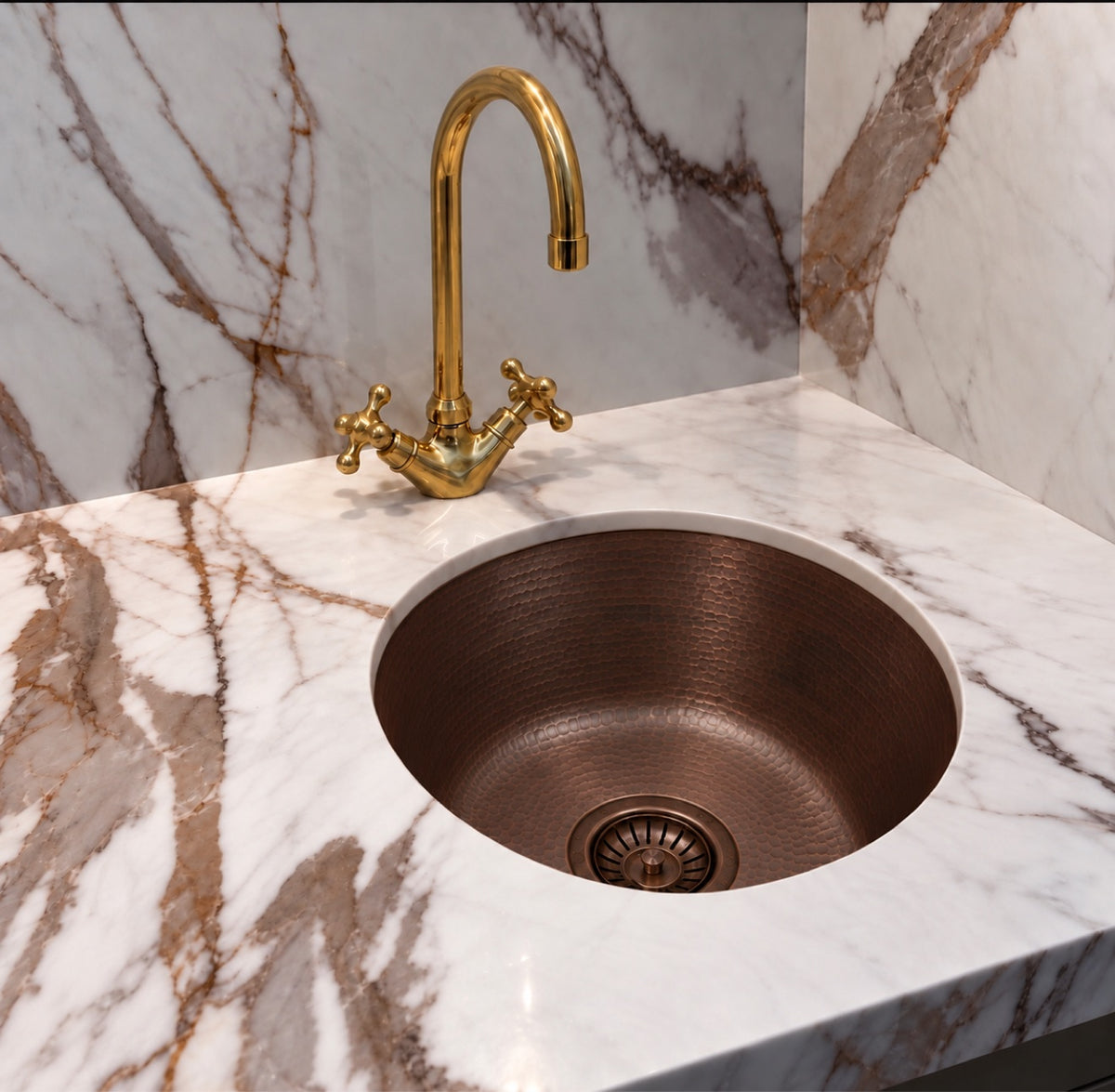 Round hammered antique copper bar sink in a marble corner bar with a brass cross handle faucet and matching strainer